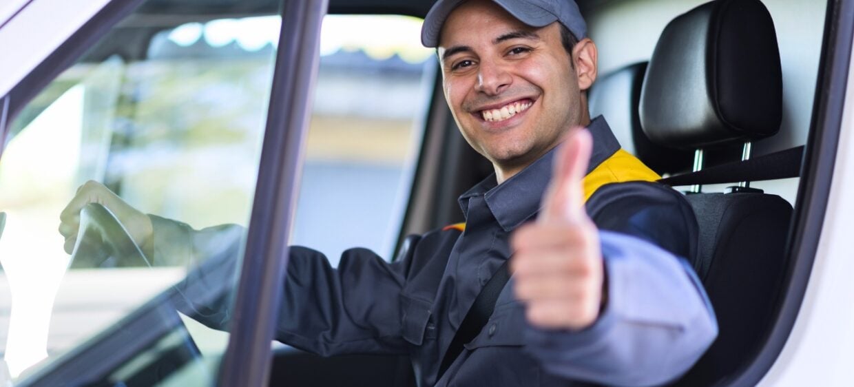 Joven migrante sonríe con un pulgar arriba mientras sujeta el volante de una van en su nuevo trabajo gracias a tener el carnet de conducir español.