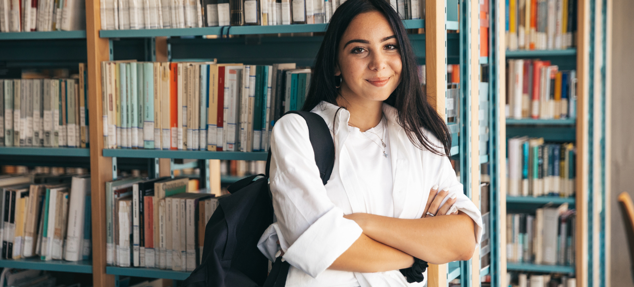 Joven con mochila sonríe en la biblioteca de la universidad donde estudia grado en España