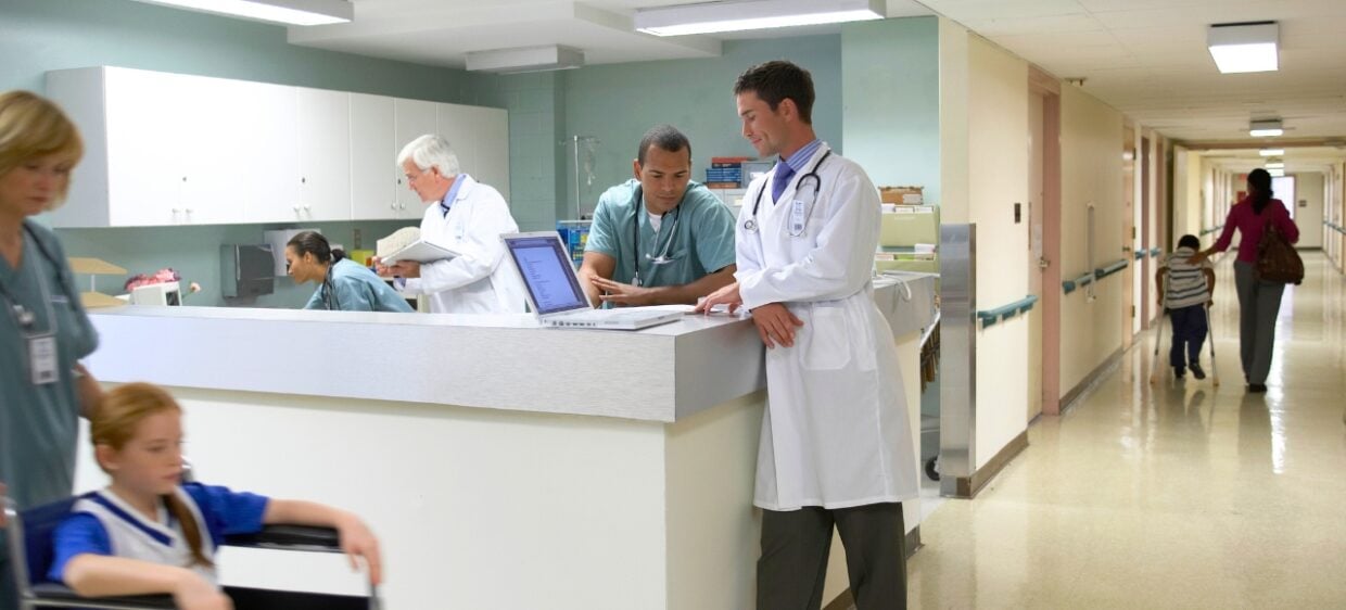 Médicos revisan un ordenador en un centro de salud mientras pacientes circulan por los pasillos